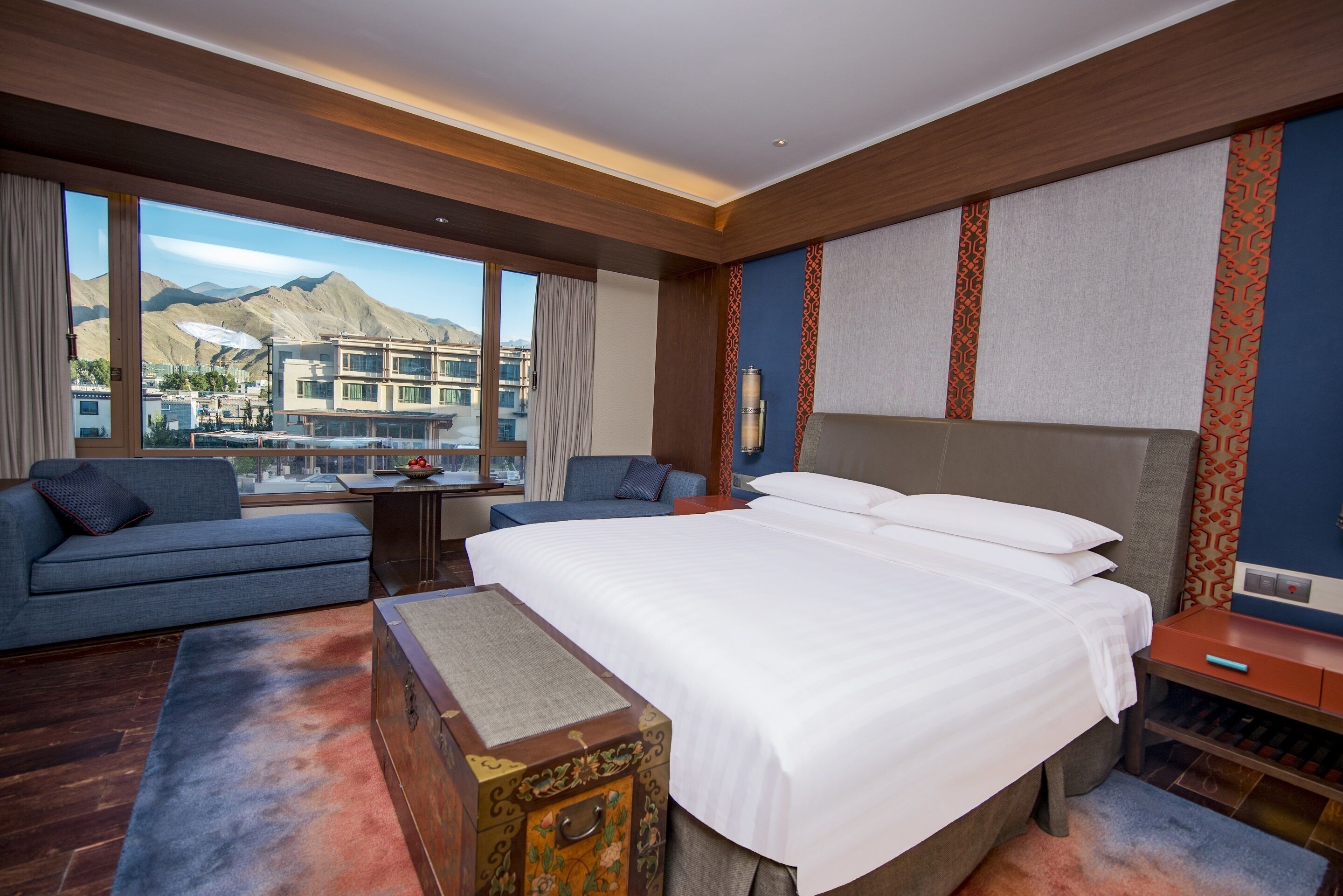 A hotel room with a large bed, a sofa, a small table, and a view of mountains and buildings.
