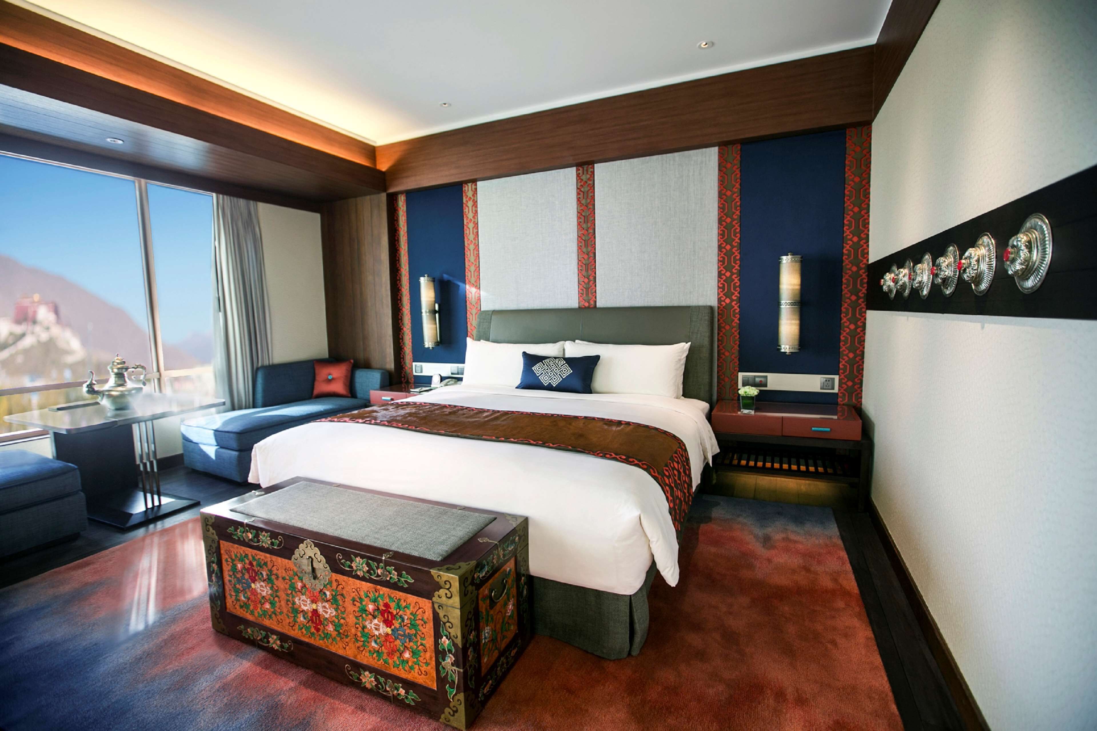 A hotel room with a large bed, a small table, a sofa, a decorative chest, and a view of mountains.