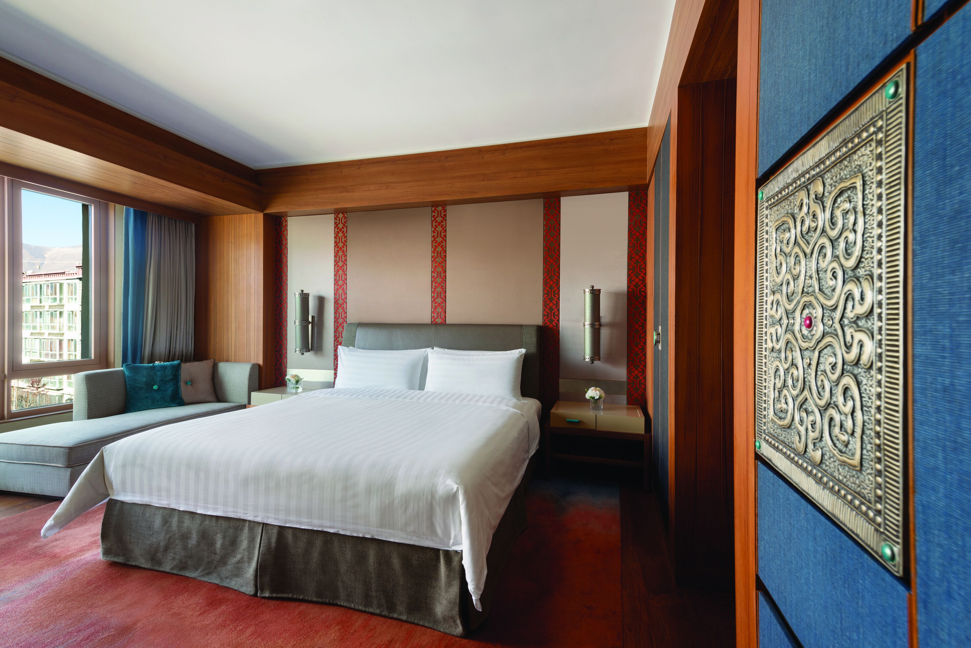 Una habitación de hotel con una cama grande, una zona de estar con sofá y sillón, una mesita de noche con lámpara y un panel decorativo en la pared.