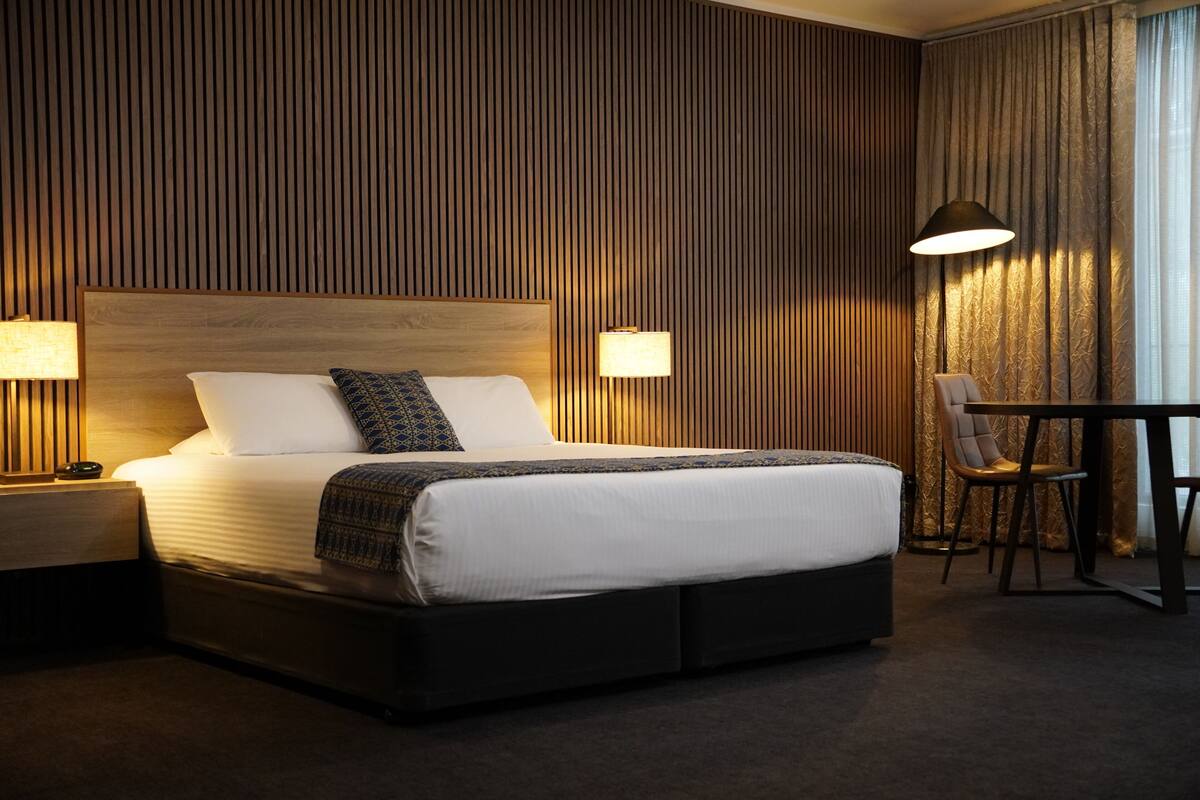 A hotel room with a large bed, two bedside lamps, a table with a chair, and a curtain with a pattern.