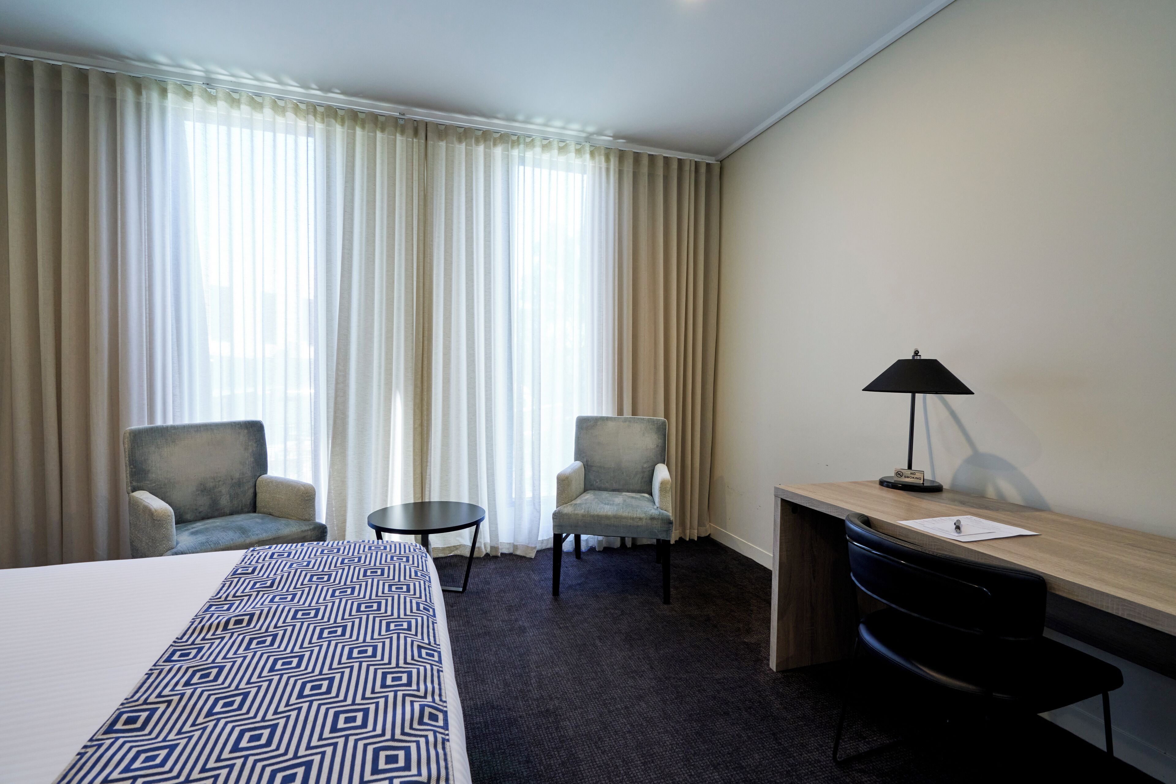 Une chambre d’hôtel avec un lit, deux fauteuils, un petit bureau avec une lampe et une vue à travers des rideaux légers.