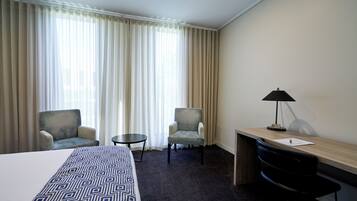 Une chambre d’hôtel avec un lit, deux fauteuils, un petit bureau avec une lampe et une vue à travers des rideaux légers.
