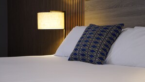 Una cama bien hecha con ropa blanca y un cojín estampado, una lámpara fijada a la pared y un cabecero de madera.
