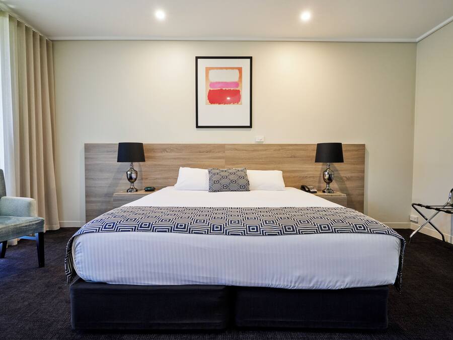 Habitación de hotel con una cama grande, dos mesitas de noche con lámparas, una silla y una obra de arte enmarcada en la pared.
