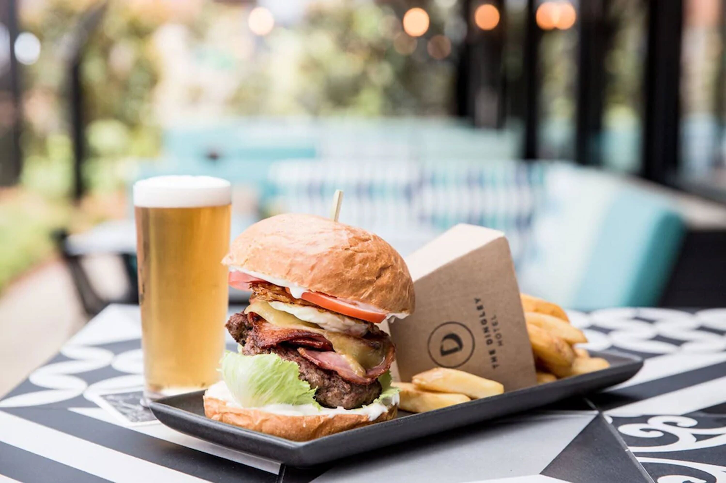 Una bandeja con una hamburguesa, papas fritas y una cerveza, con una piscina al fondo.