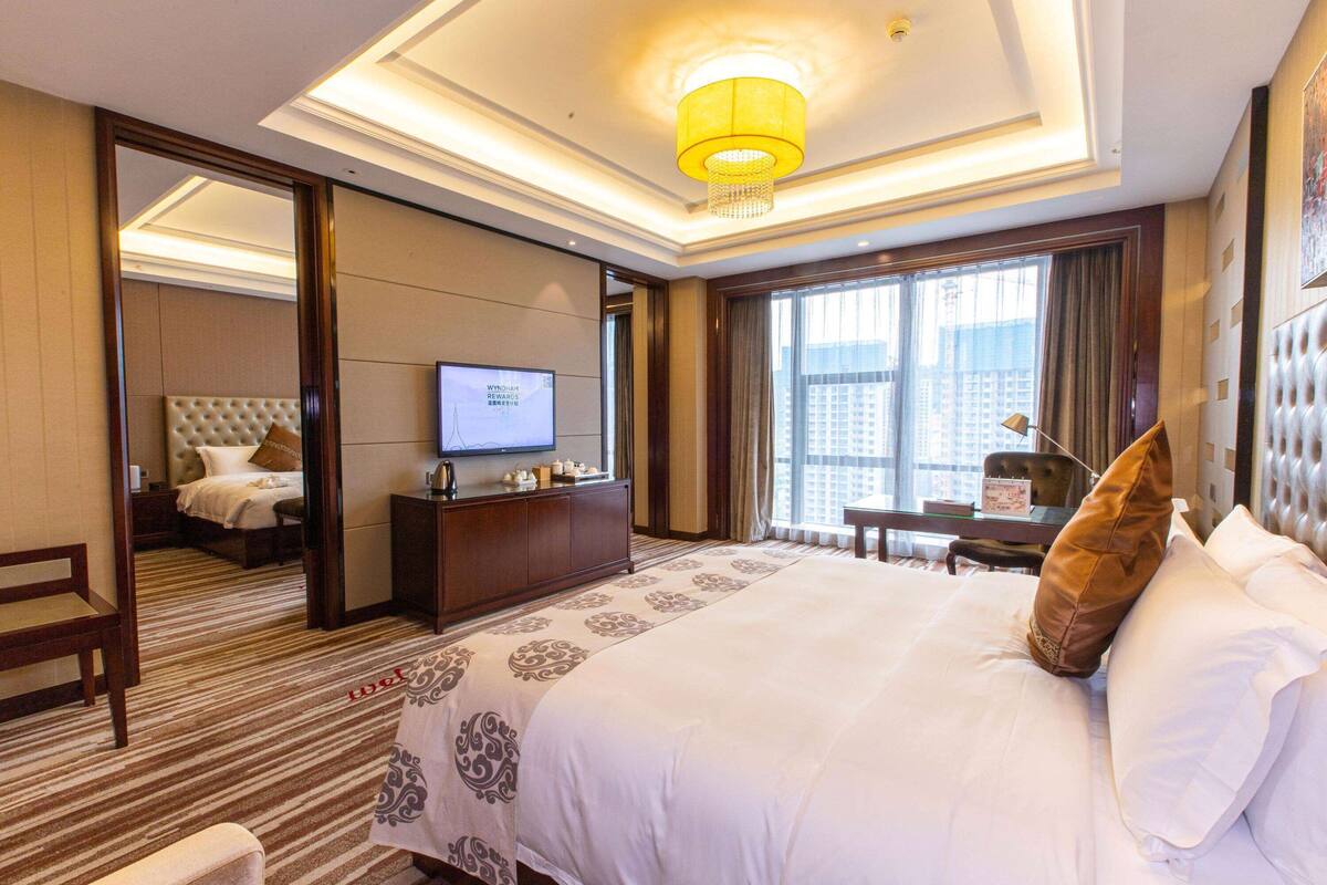 A hotel room with a large bed, a television, a desk with a lamp, and a view of the city through the window.
