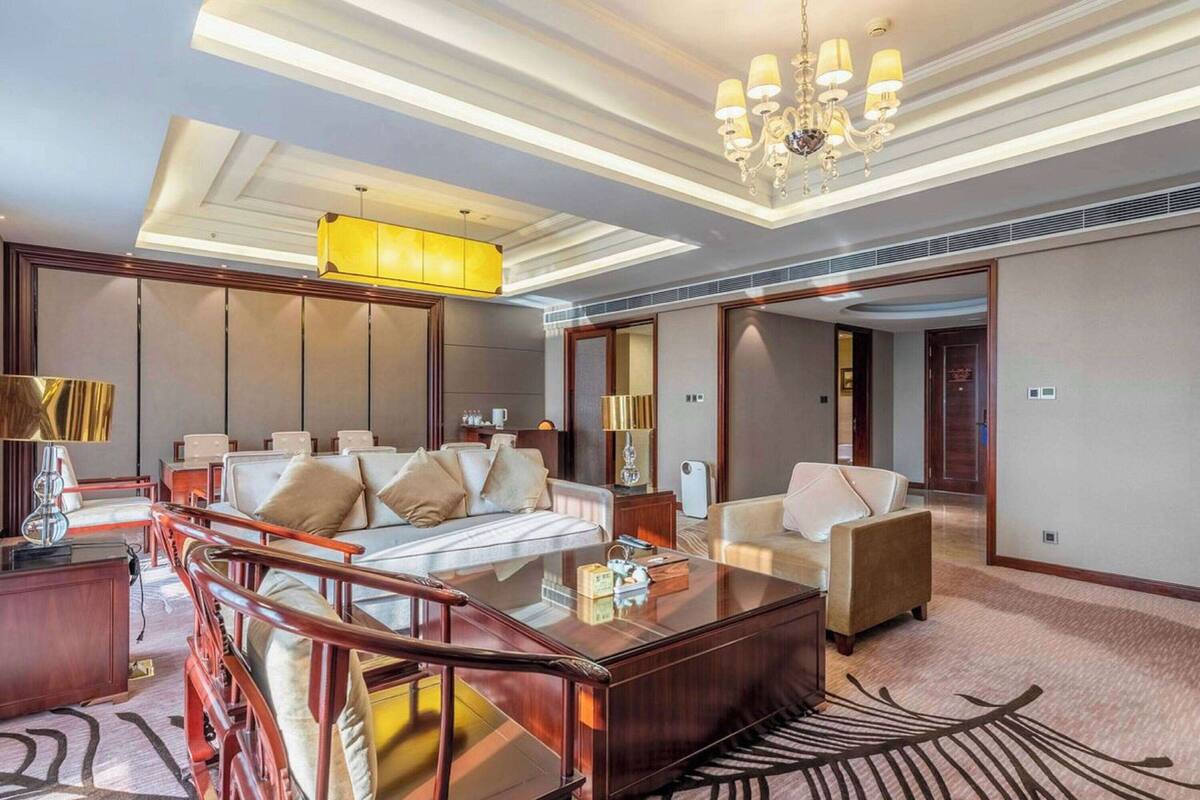 A spacious hotel room with a large bed, a wooden desk, a glass coffee table, and a seating area with a sofa and armchairs.