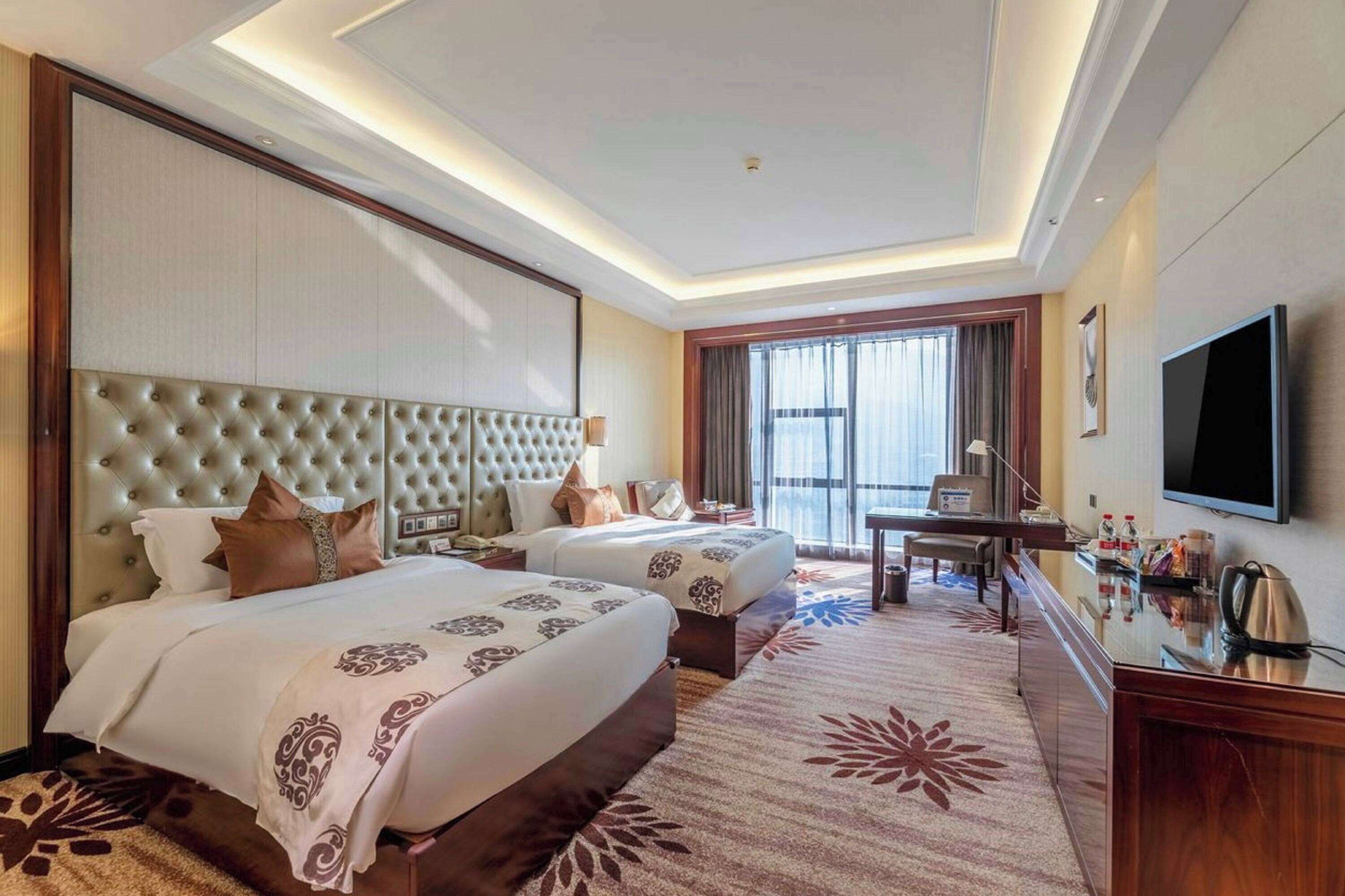 A hotel room with two beds, a desk, a television, and a large window.