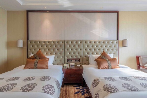 A hotel room with two beds, a large padded headboard, a bedside table with a phone and lamp, and a carpeted floor.
