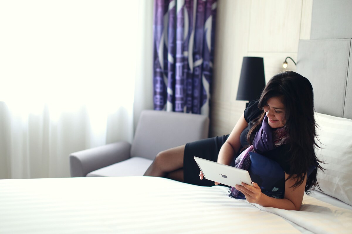A person lying on a bed in a hotel room, using a tablet.