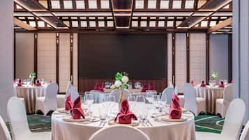 Un salón de banquetes con mesas redondas, sillas blancas y una gran pantalla al frente.
