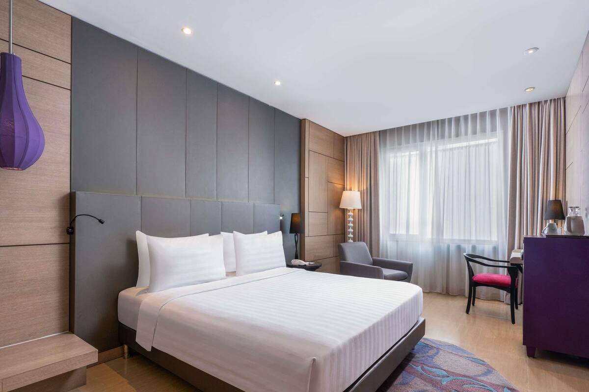A modern hotel room with a large bed, a desk, a chair, and a lamp.