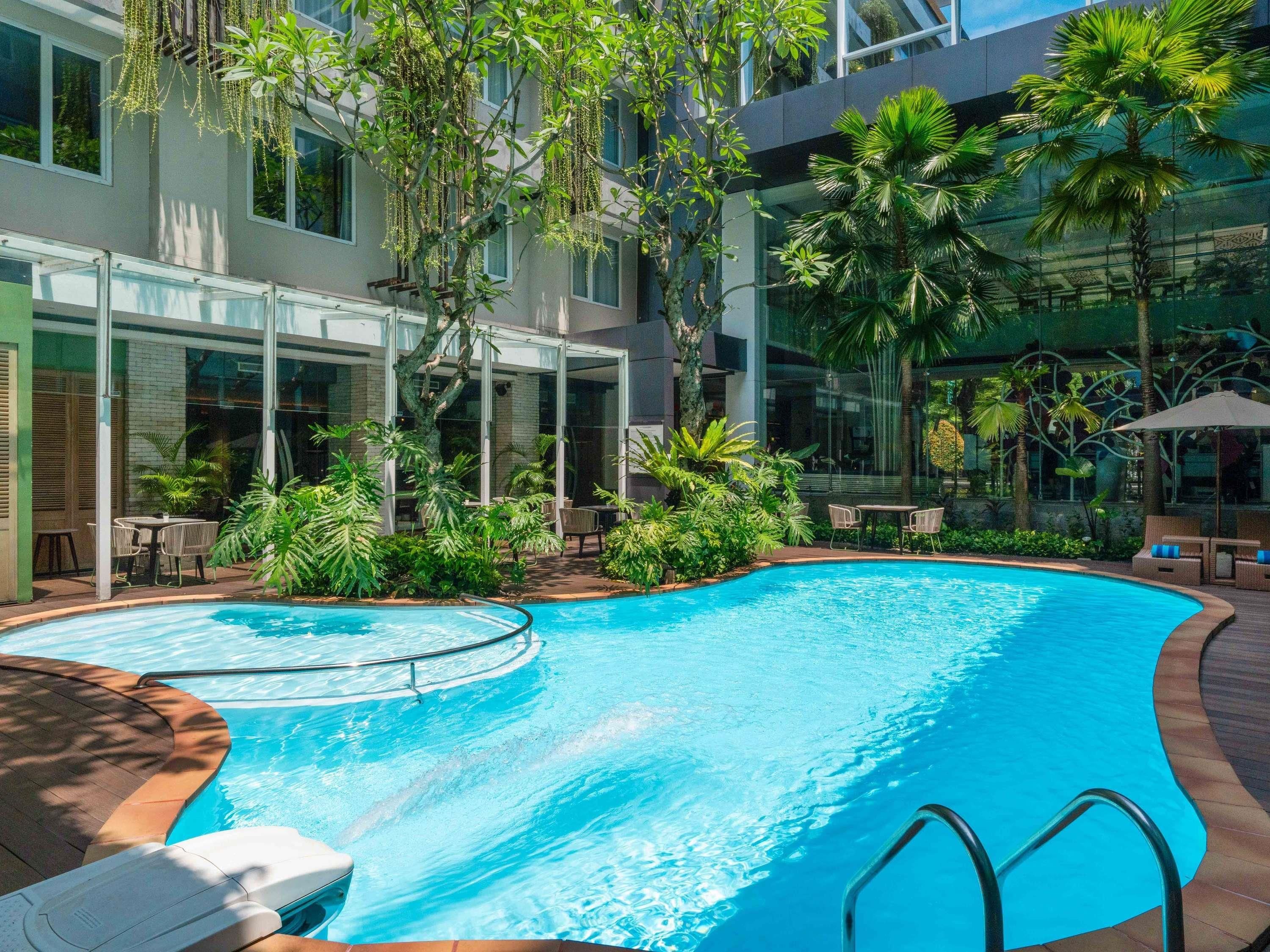 Una piscina con cerramiento de vidrio, rodeada de exuberante vegetación y mobiliario de exterior.