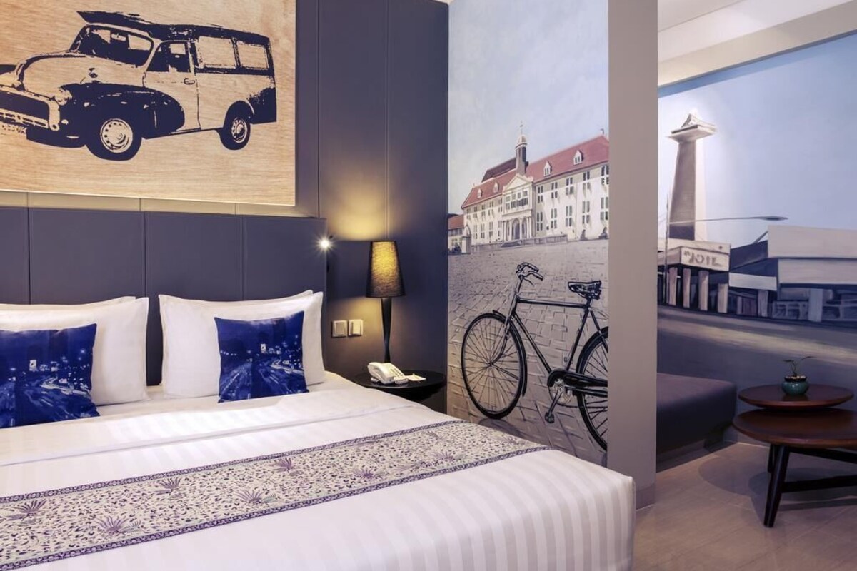A hotel room with a large bed, a nightstand, a lamp, and a wall with a bicycle and a building.