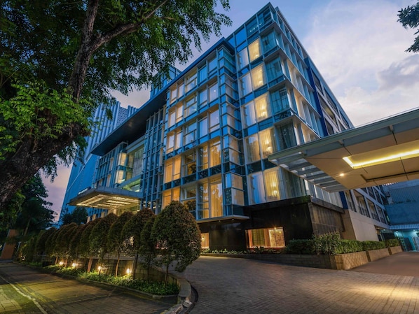 A modern building with a glass facade, a covered entrance, and a landscaped area with trees. - Mercure Jakarta Sabang (Jakarta)