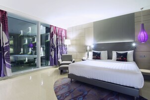 A modern hotel room with a large bed, a seating area, and a glass wall offering a view of the interior.