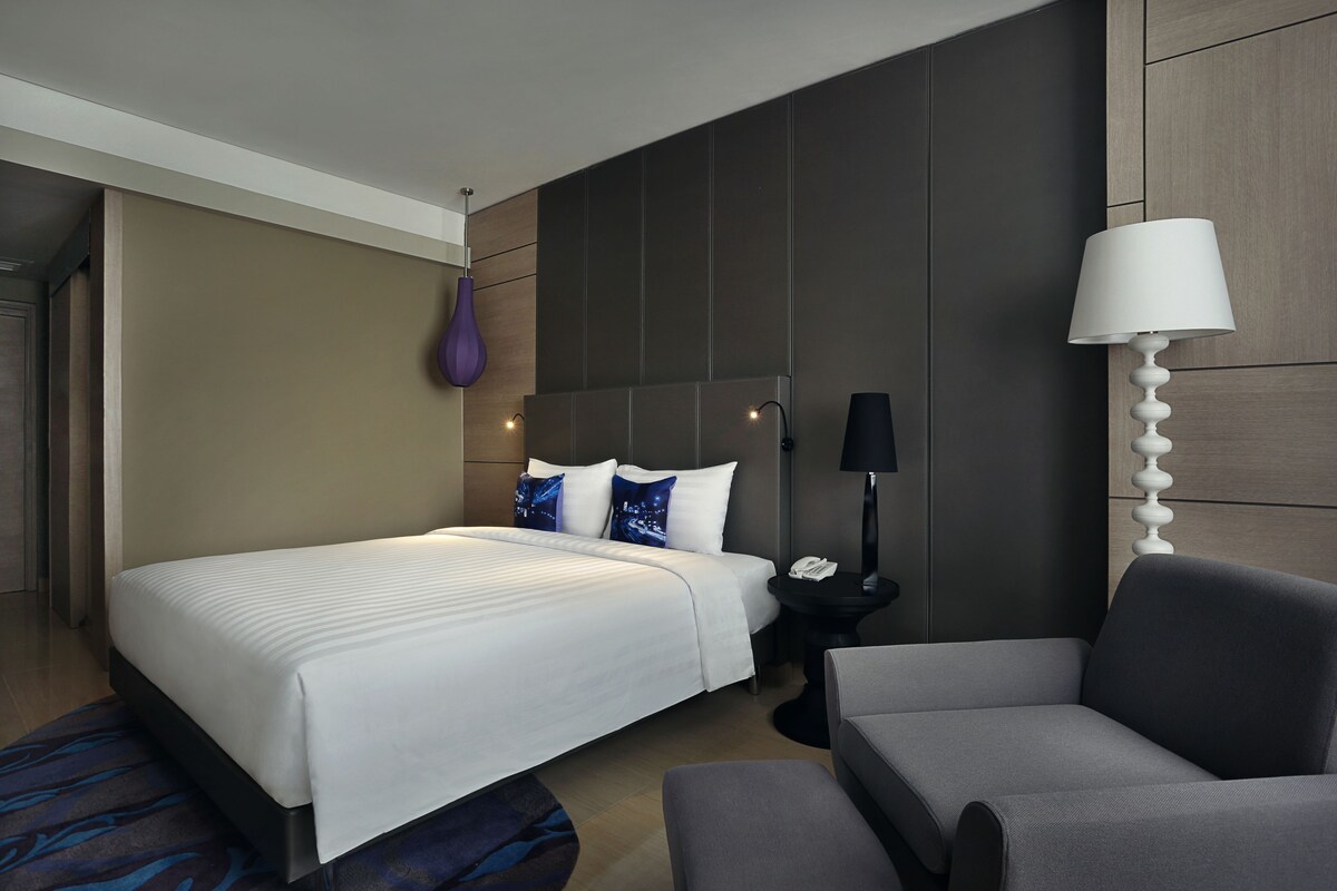 A modern hotel room with a large bed, a gray armchair, a small side table with a lamp, and a purple vase on the wall.