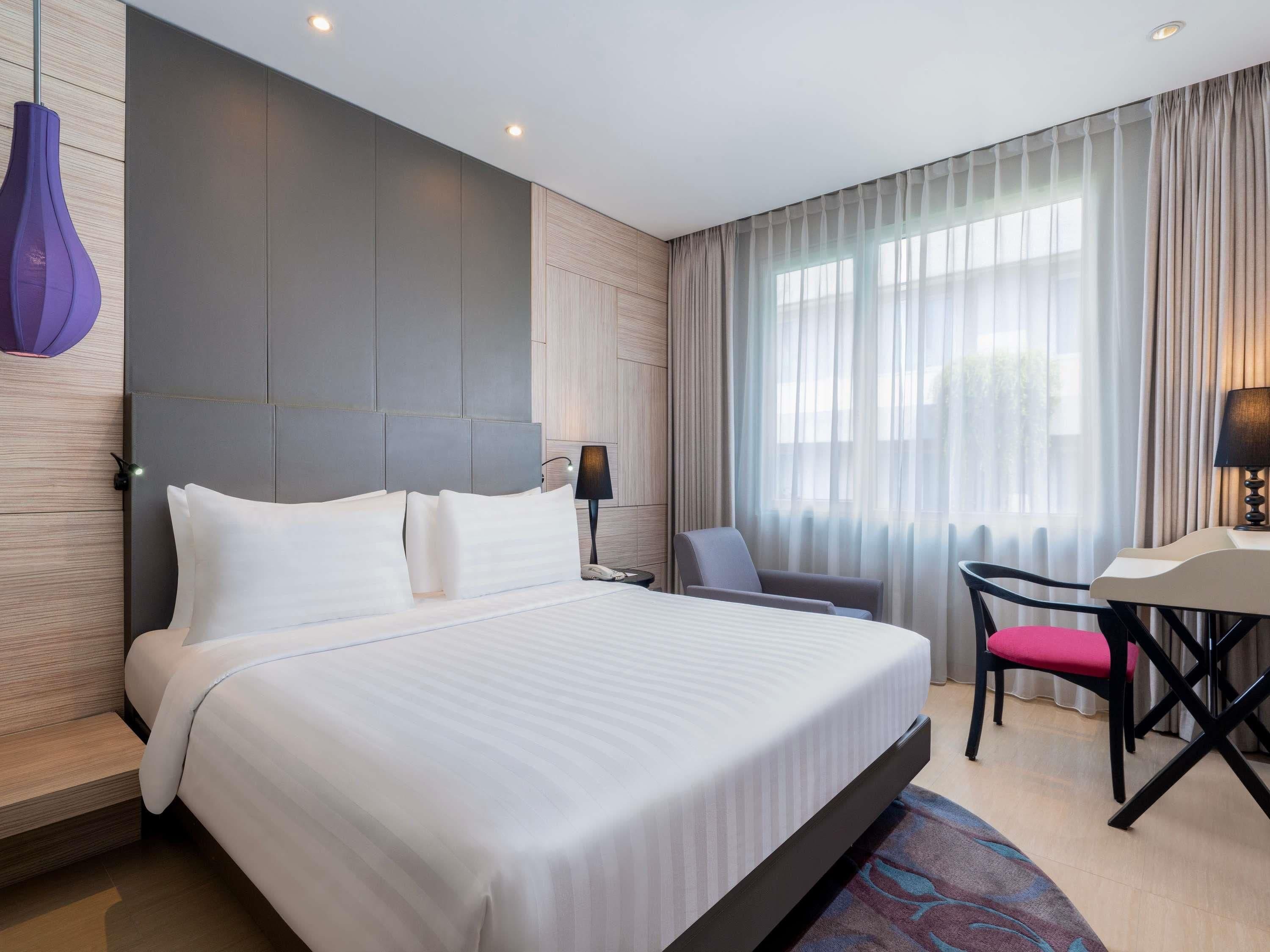 A hotel room with a large bed, a desk with a chair, a lamp, and a window with curtains.