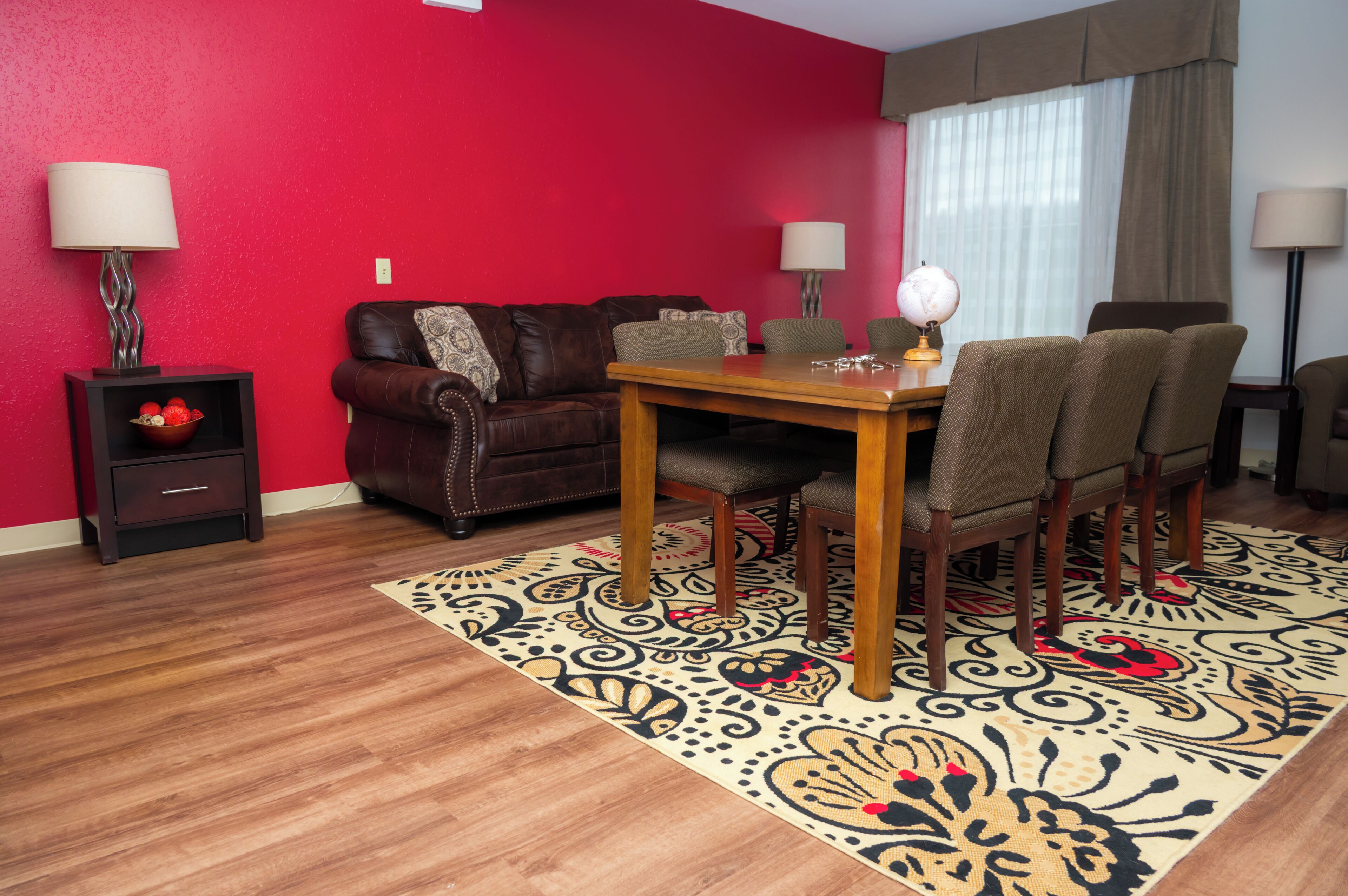 A living room with a red wall, a brown leather sofa, a wooden dining table with four chairs, a patterned rug, and two side tables with lamps and decorative bowls.