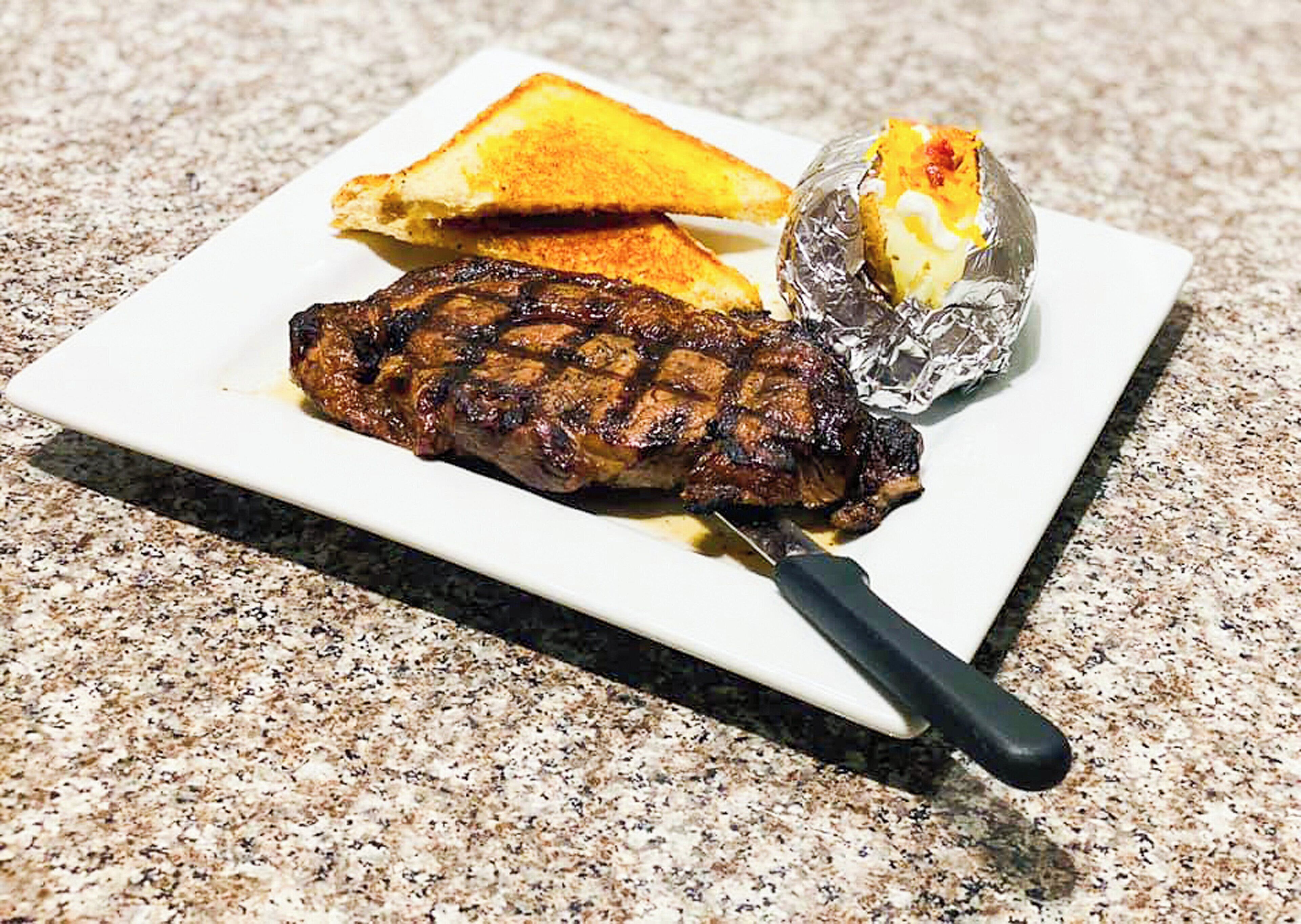 A grilled steak with a knife, a grilled sandwich, and a foil-wrapped item on a white plate.