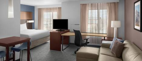 Habitación de hotel con cama, escritorio, computadora, silla, sofá y ventanas amplias.
