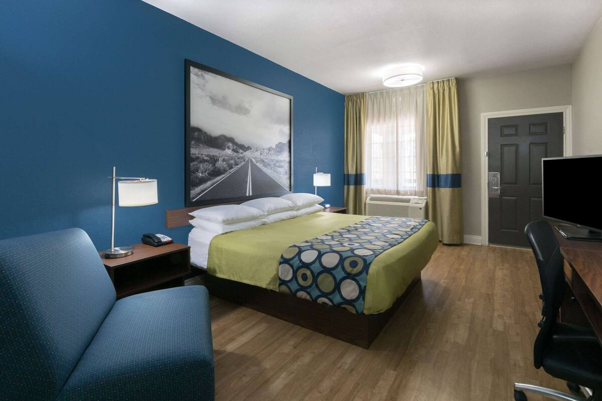 A hotel room with a bed, a desk with a computer, a chair, a nightstand, a lamp, and a large framed picture on the wall.