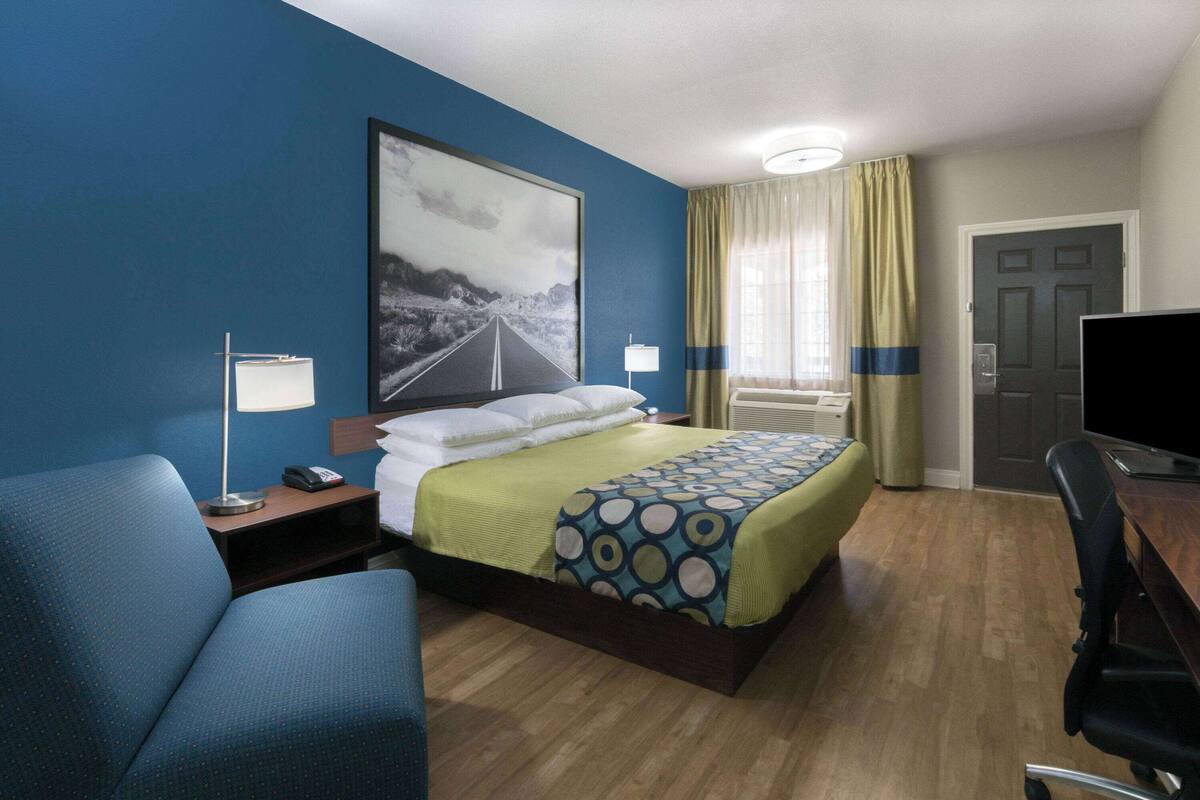 A hotel room with a bed, a desk, a chair, a lamp, a TV, and a painting on the wall.