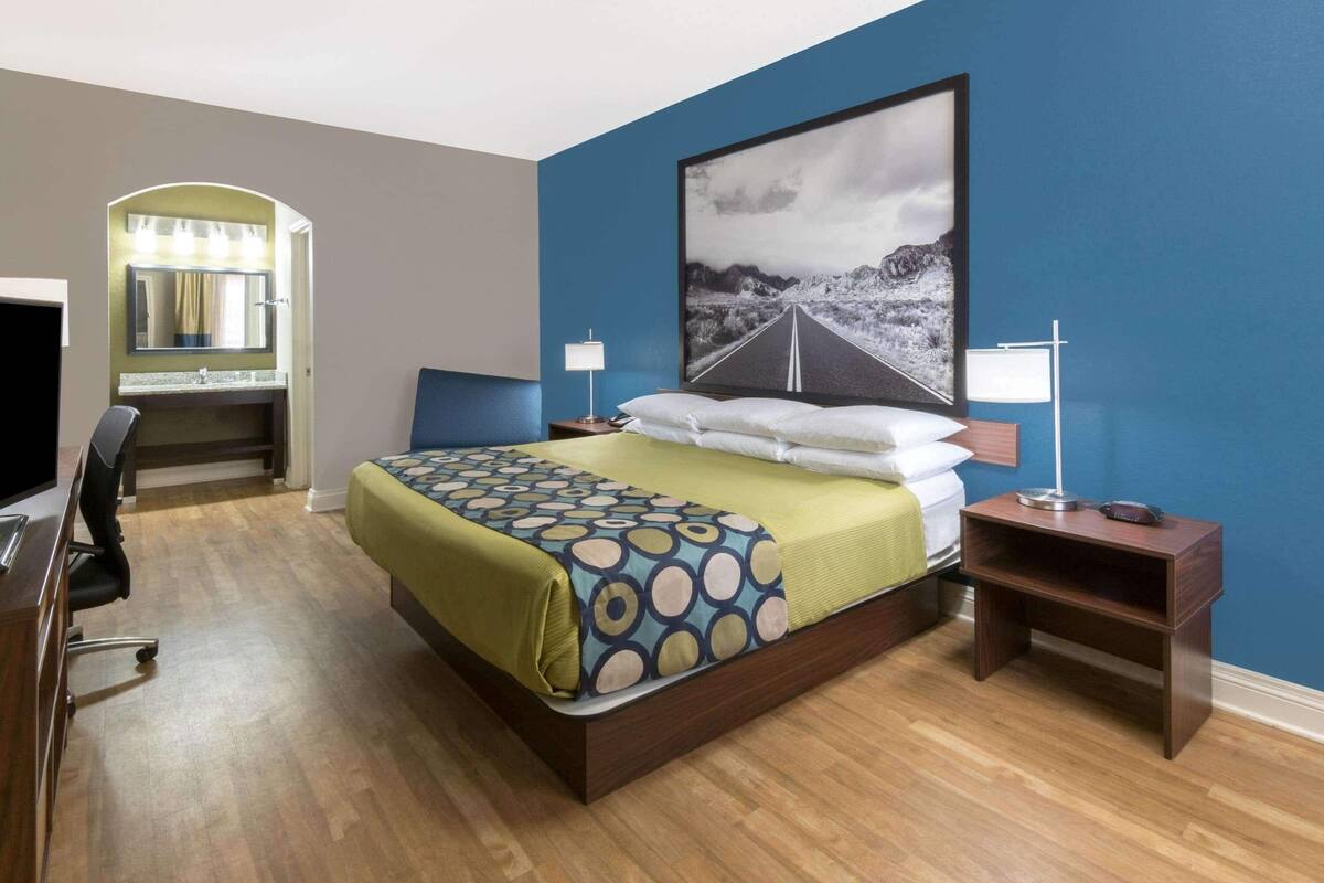 A hotel room with a bed, a desk, a chair, a TV, a nightstand, and a painting on the wall.