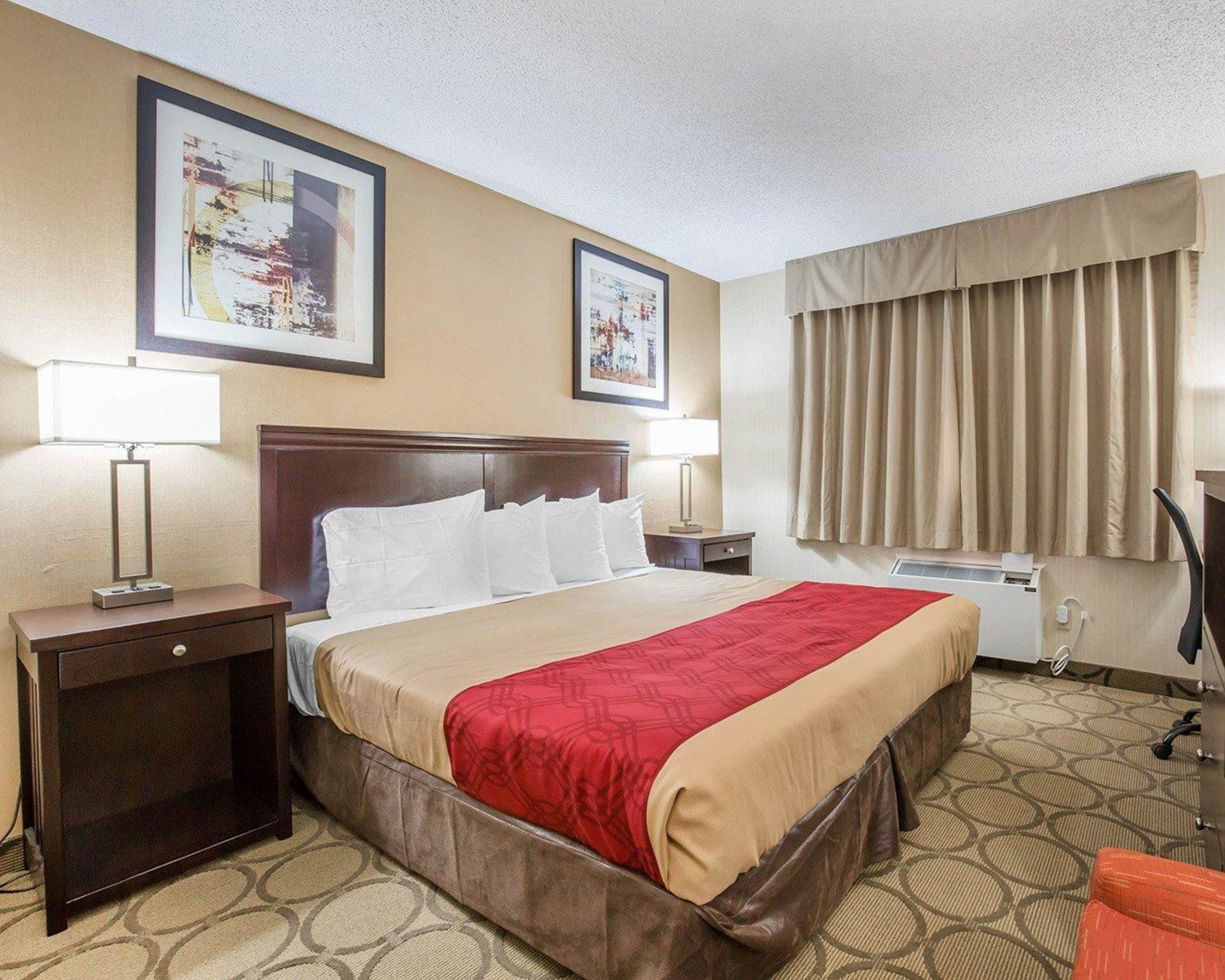 A hotel room with a large bed, two bedside tables with lamps, a window with curtains, and two framed pictures on the wall.