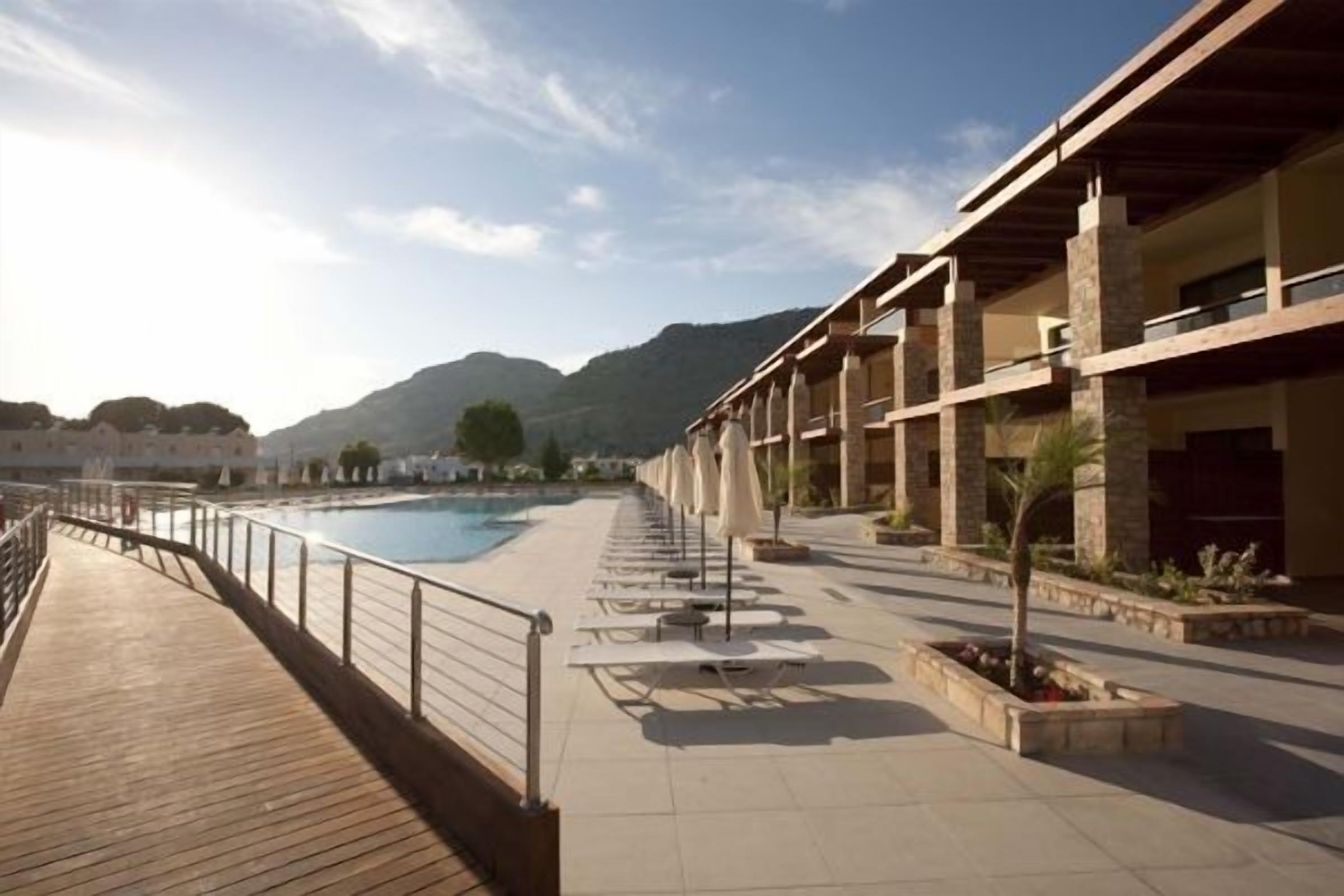 A resort pool area with a wooden deck, lounge chairs, and a building with multiple balconies.