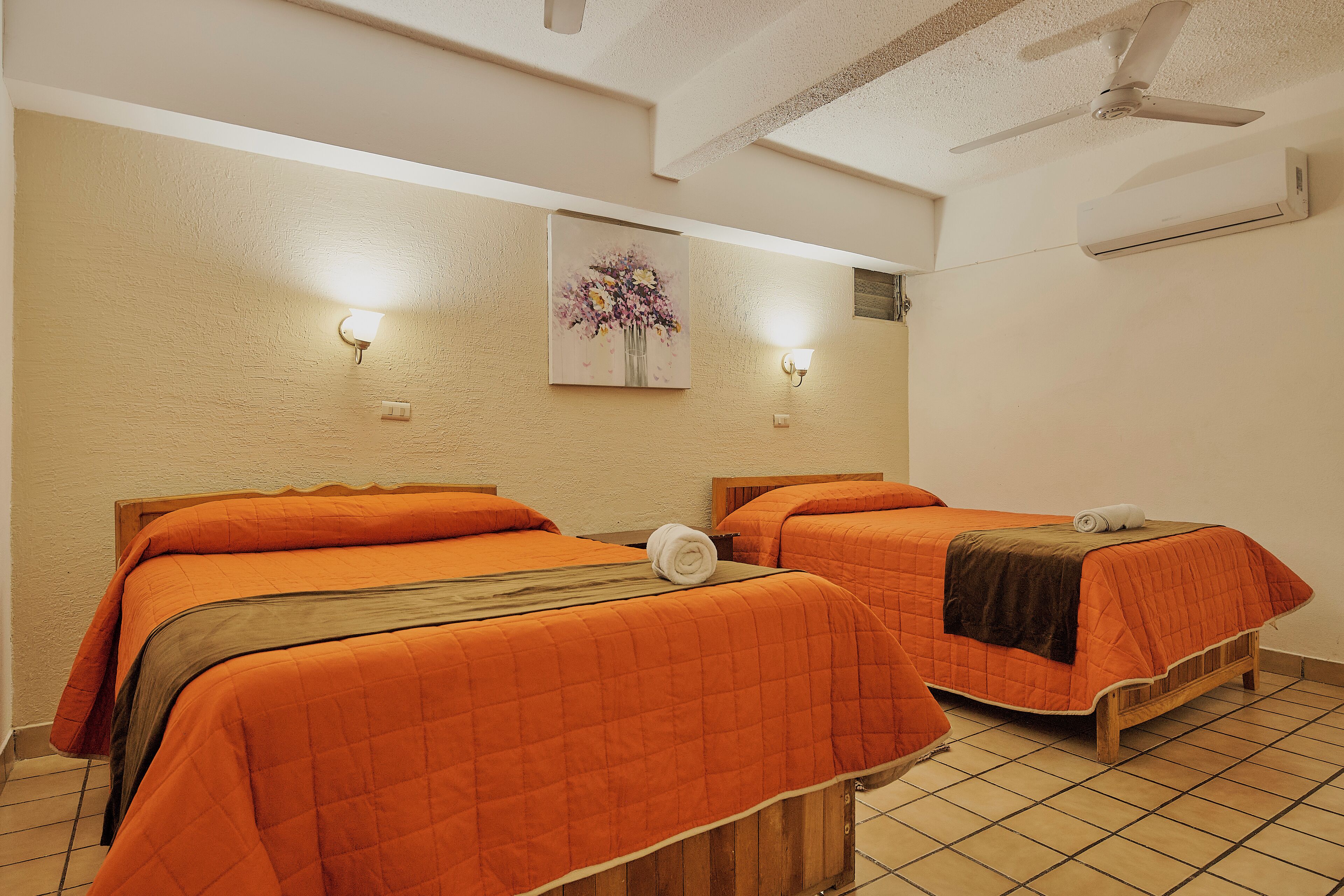 Habitación de hotel con dos camas, cada una con colcha naranja, un cuadro en la pared y un ventilador de techo.