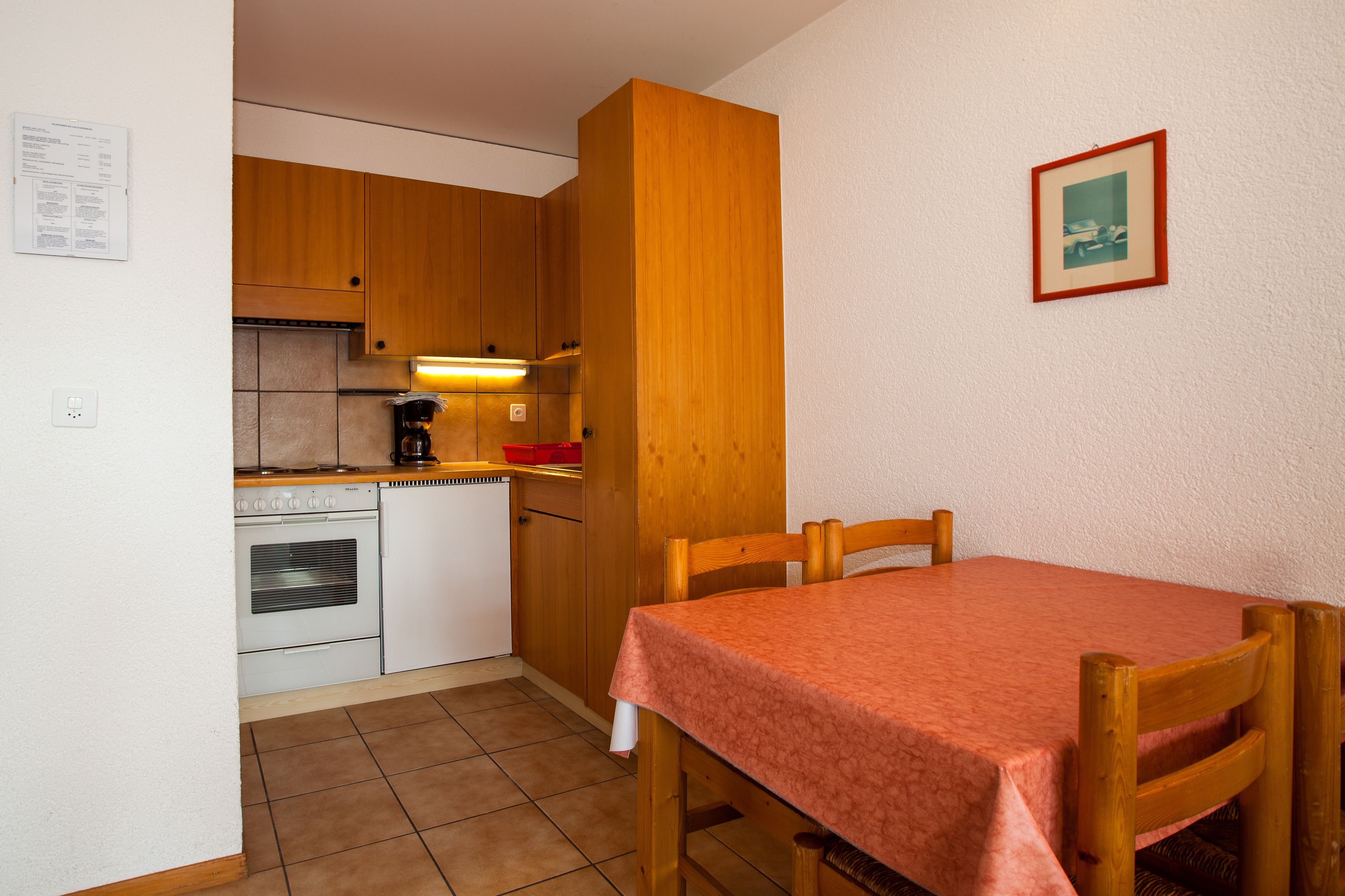 Une cuisine compacte avec des meubles en bois, un four encastré et une table à manger recouverte d’une nappe rouge.