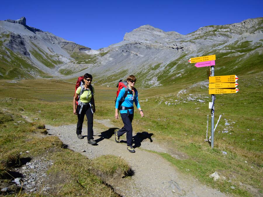 Two hikers on a mountain trail with a directional signpost.