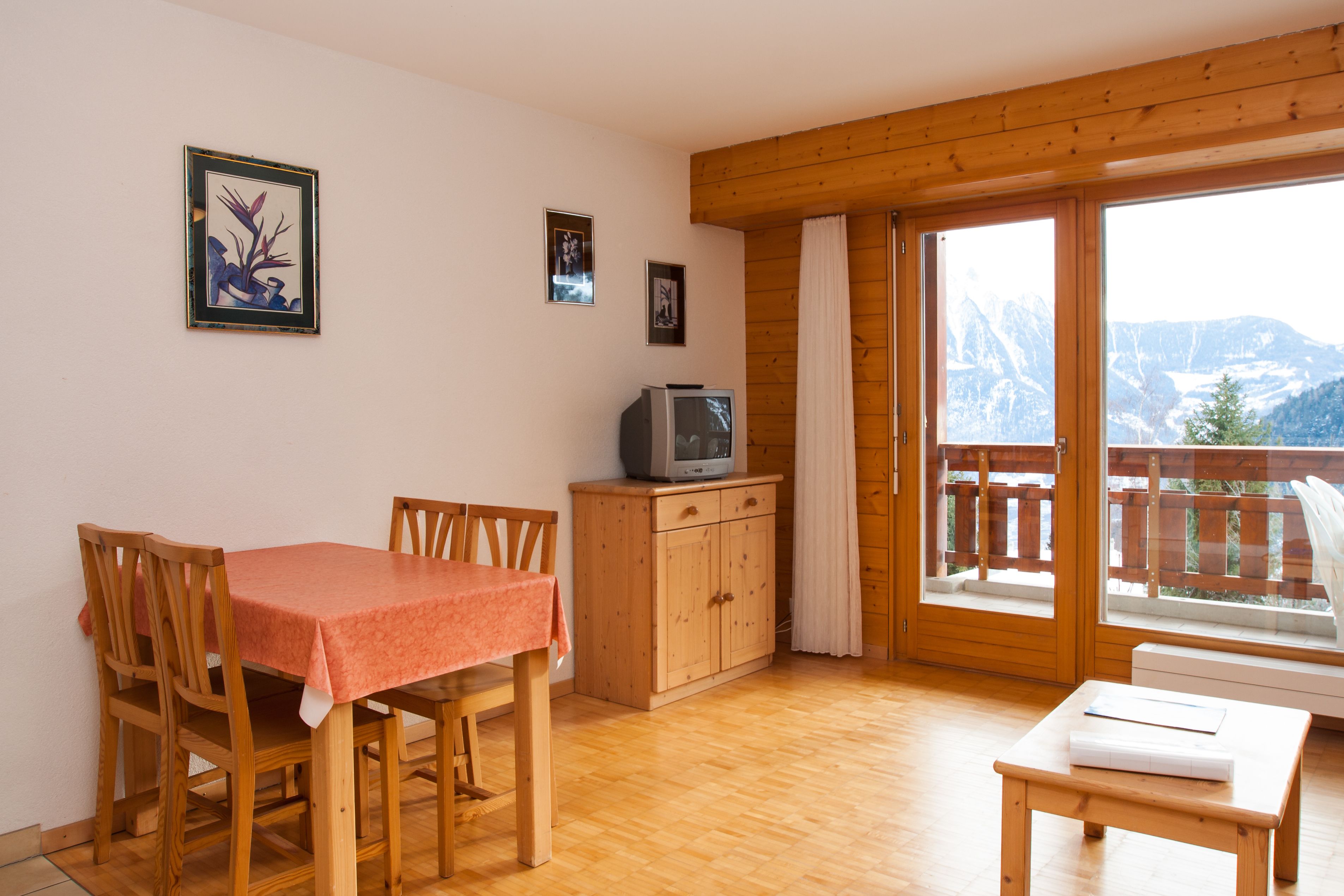 Un comedor con una mesa y sillas, un televisor sobre un mueble de madera, y un balcón con vistas a montañas.