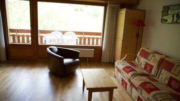 A living room with wooden flooring, a sofa, a chair, a table, and a window with a view of outdoor furniture.