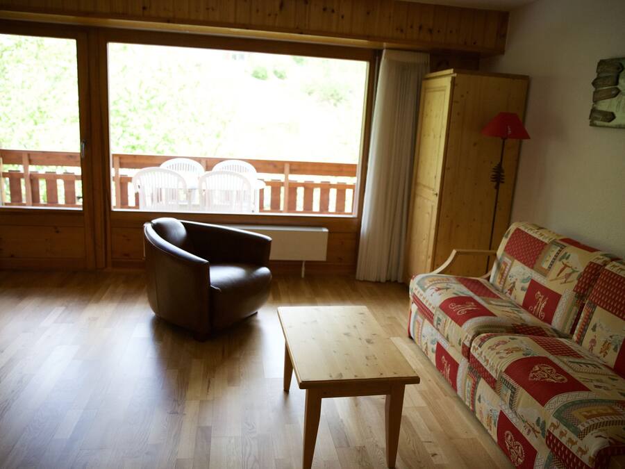A living room with wooden flooring, a sofa, a chair, a table, and a window with a view of outdoor furniture.