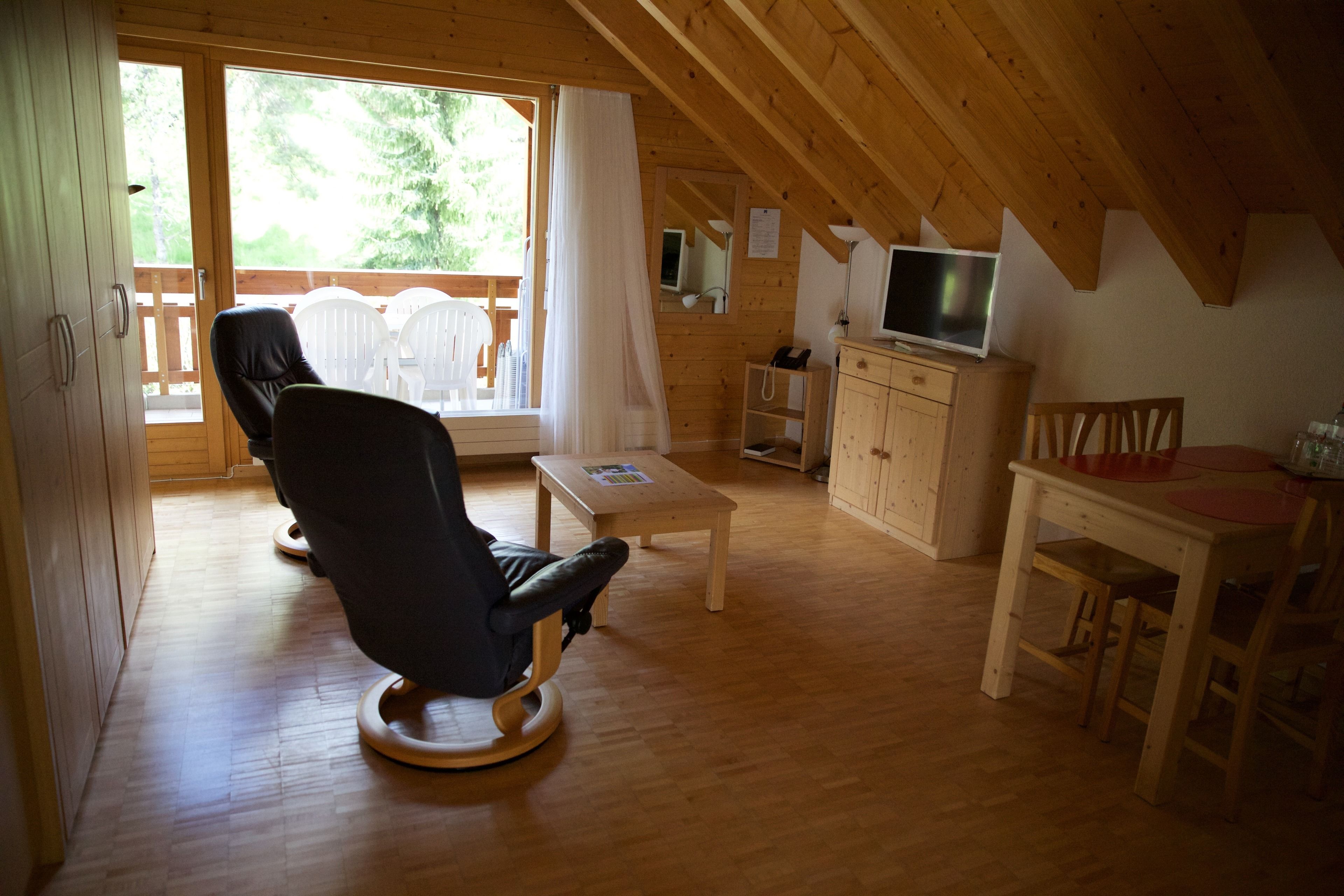 Una cabaña acogedora con interior de madera, un sillón reclinable cómodo, una mesita, un televisor y un balcón con sillas blancas.