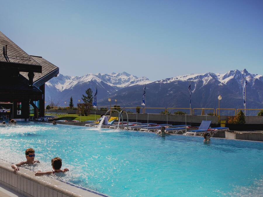 Una piscina con agua cristalina azul, rodeada de sillas de descanso y vistas a montañas nevadas.