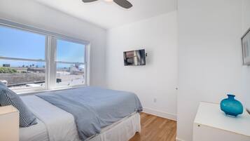 A bedroom with a bed, a ceiling fan, a TV, and a window with a view of buildings.