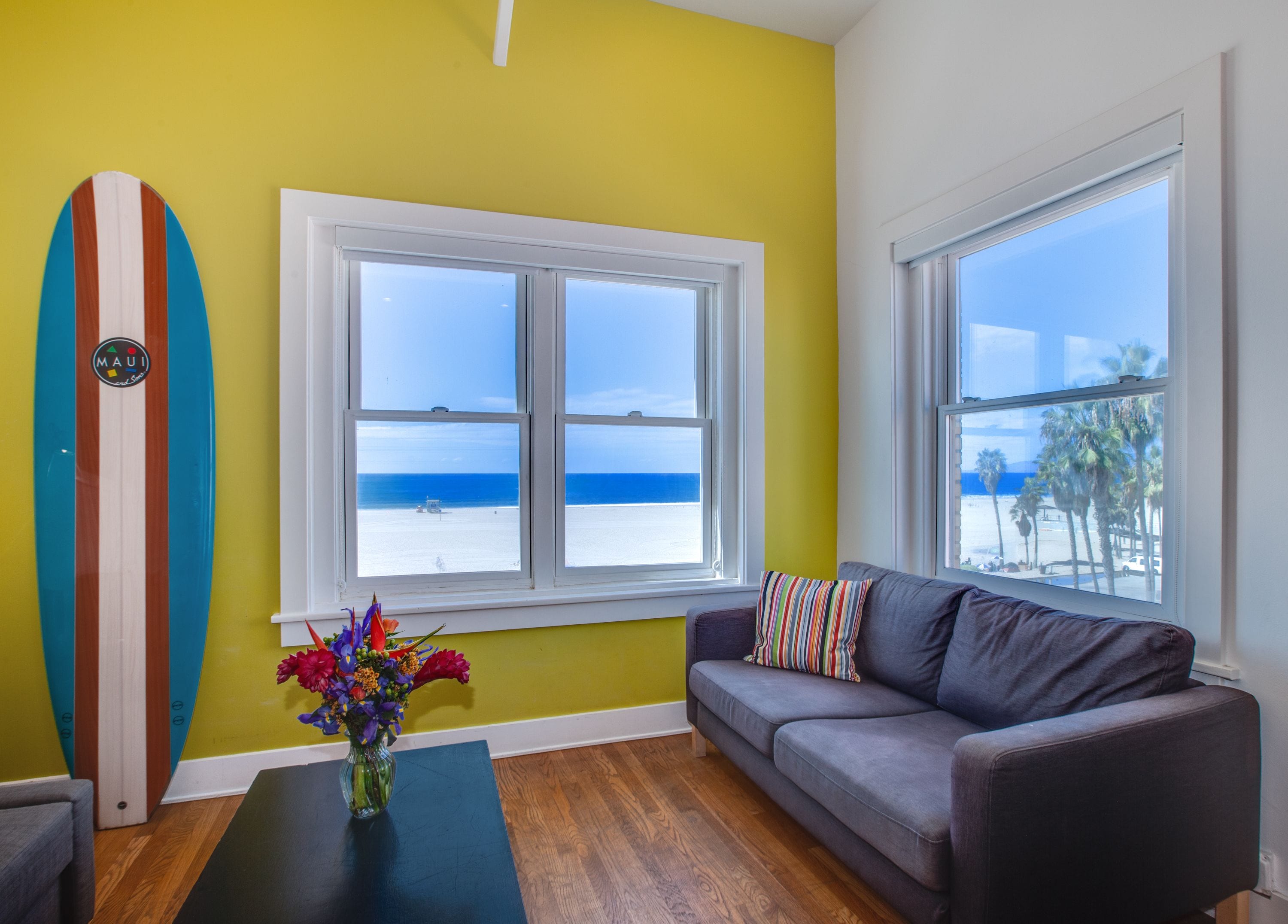 Quarto com uma janela grande que oferece vista para a praia, um prancha de surf na parede, um sofá cinzento e um vaso com flores sobre uma mesa.