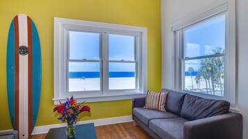 Quarto com uma janela grande que oferece vista para a praia, um prancha de surf na parede, um sofá cinzento e um vaso com flores sobre uma mesa.