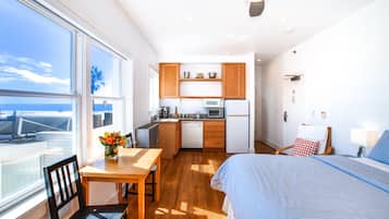 A compact hotel room with a kitchenette, a bed with a blue-striped duvet, a dining table with a chair, and a large window offering a view of the sea.