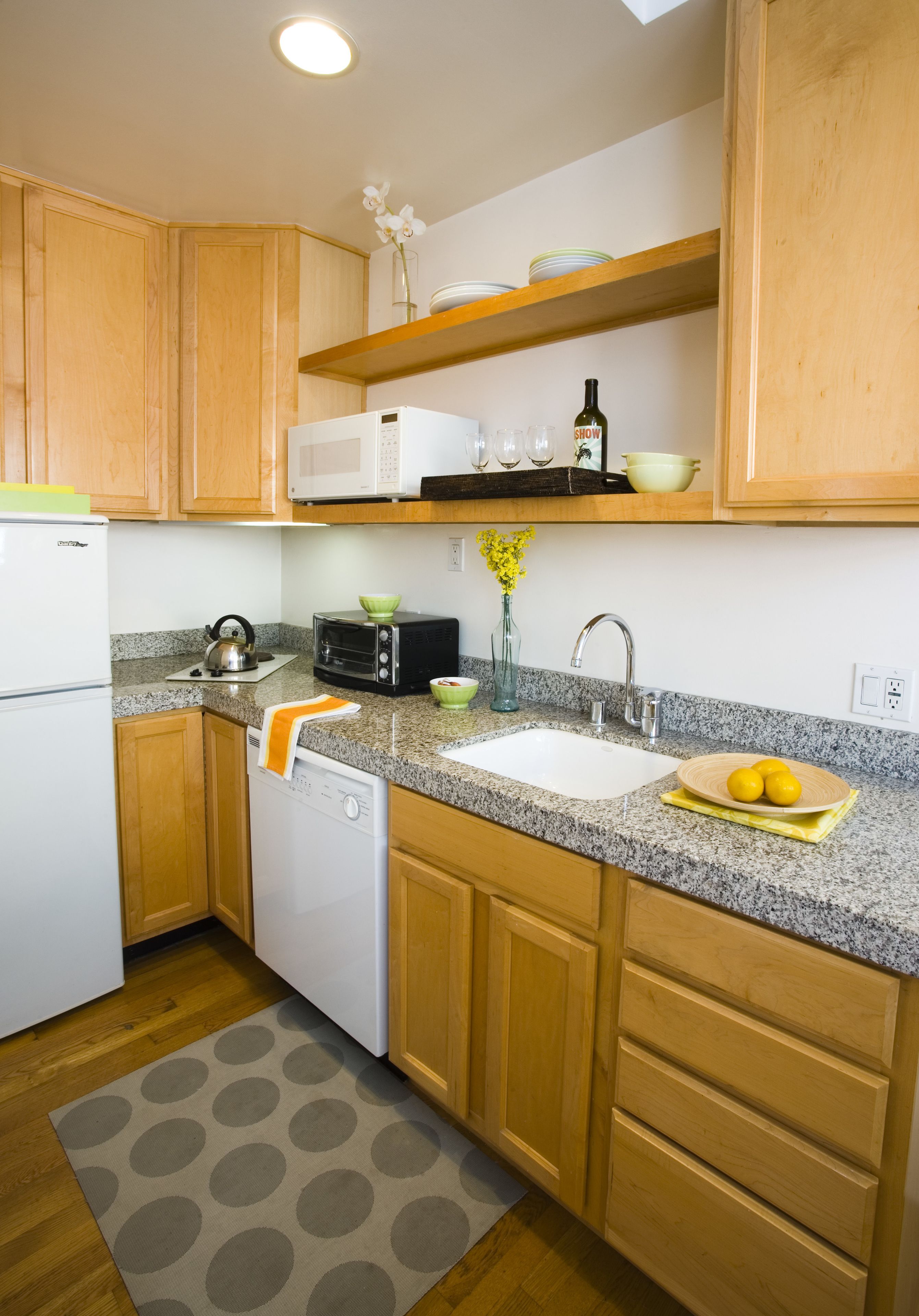 A modern kitchen with wooden cabinets, a granite countertop, a white refrigerator, a dishwasher, a microwave, a toaster, a sink, and a small shelf above the countertop.