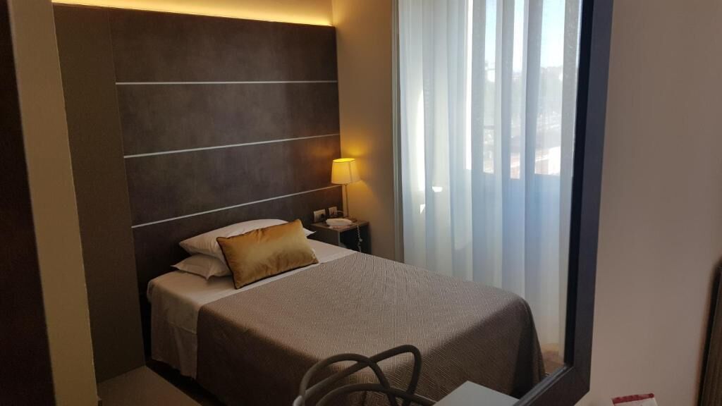 Ein Hotelzimmer mit Bett, Nachttisch, Lampe und Blick aus dem Fenster.