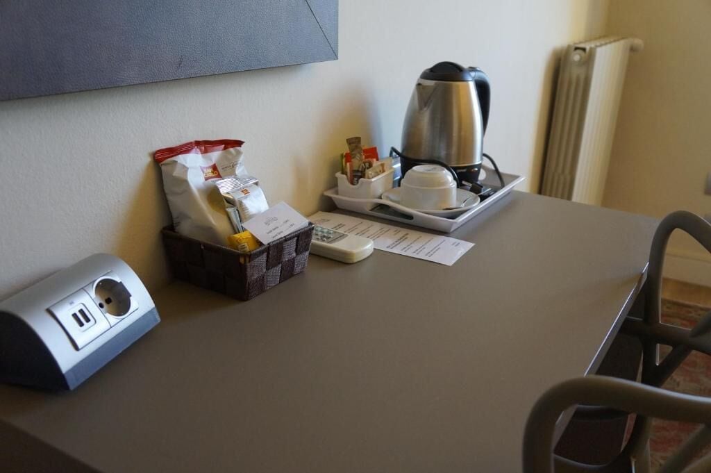 A hotel room desk with a coffee maker, a bag of snacks, a cup of coffee, a scale, and a power outlet.