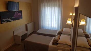 A hotel room with two beds, a TV mounted on the wall, a painting, a radiator, and a window with curtains.