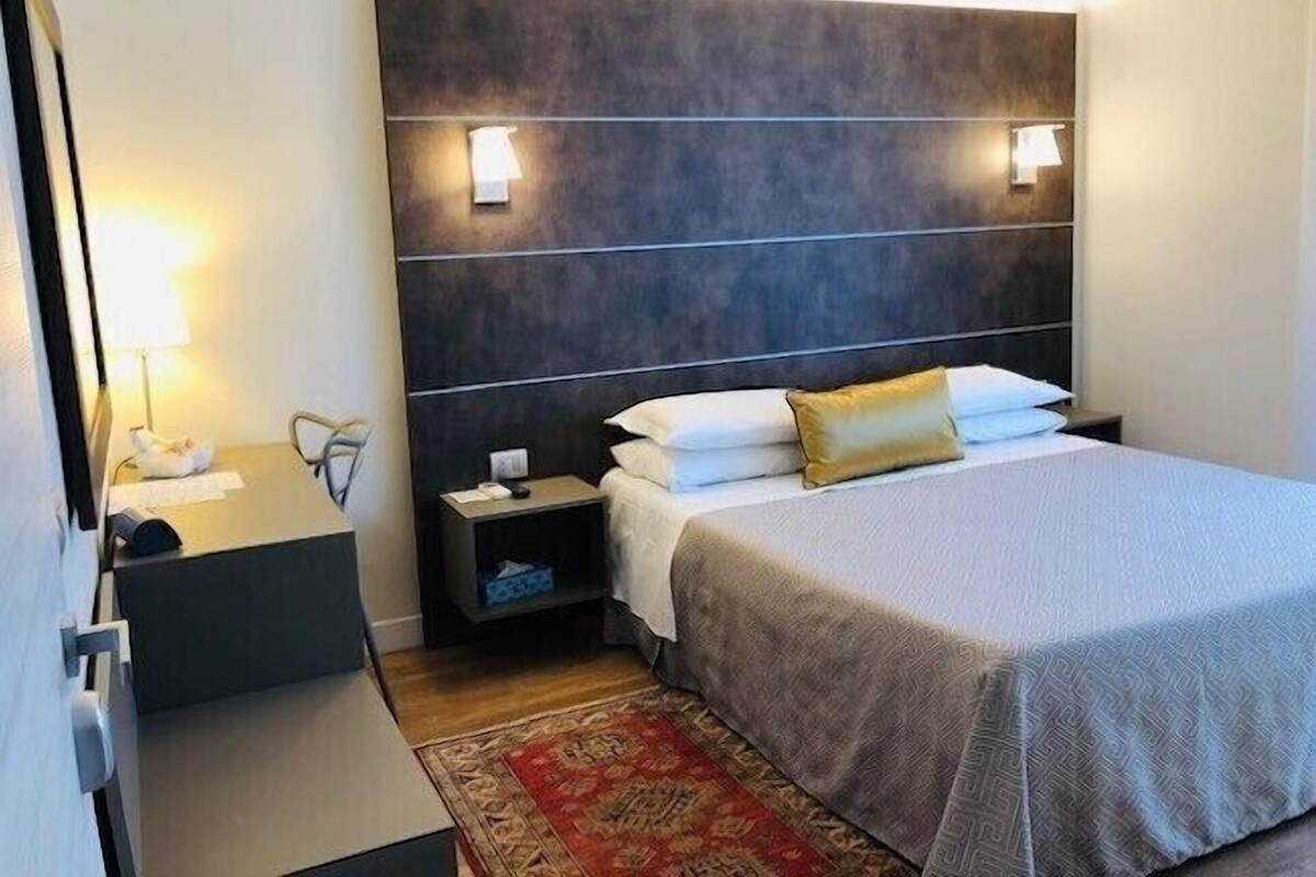 A hotel room with a bed, bedside tables, a nightstand, a lamp, and a decorative rug.