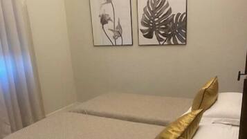Un lit double avec un oreiller blanc et doré, une table de chevet avec une télécommande, et deux œuvres d’art encadrées accrochées au mur.