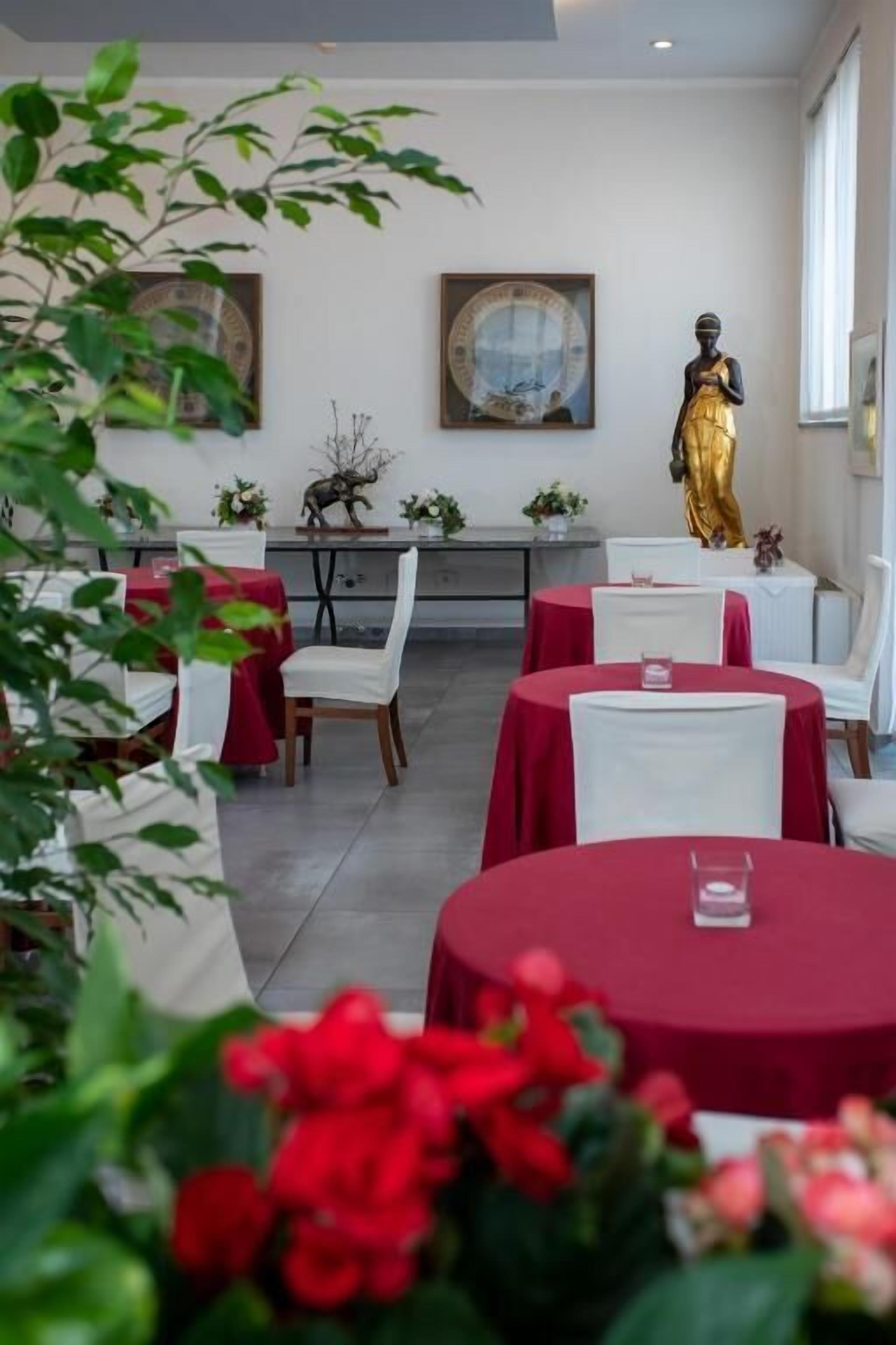 Zona pranzo con tavoli rotondi, sedie bianche e tovaglie rosse. Sui tavoli ci sono oggetti decorativi e una statua sullo sfondo.