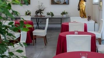 Zona pranzo con tavoli rotondi, sedie bianche e tovaglie rosse. Sui tavoli ci sono oggetti decorativi e una statua sullo sfondo.